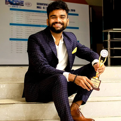 Smiling man in a dark checkered suit sits on stairs, holding a trophy with a gold cup.