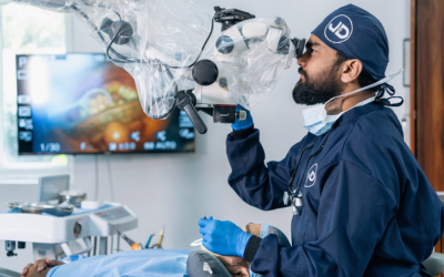 Surgeon in navy scrubs and cap uses a surgical microscope during an operation, with a monitor displaying medical images in the background.