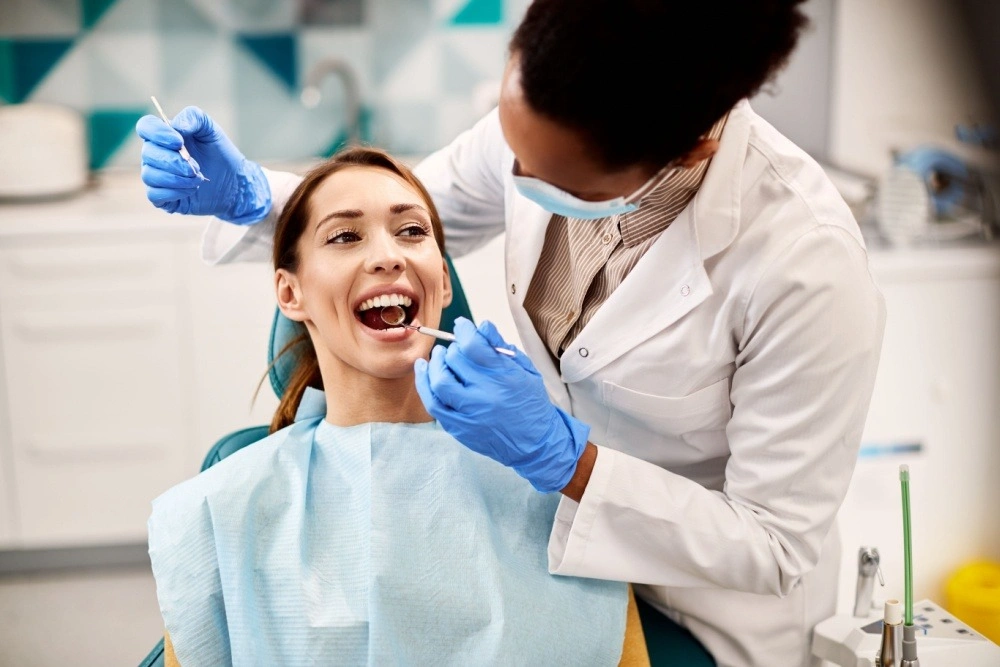 Dr_Jaydev_Dental_treatment | Jaydev Dental Dentist in gloves and mask uses dental tools to examine a woman's open mouth in a clinic setting.