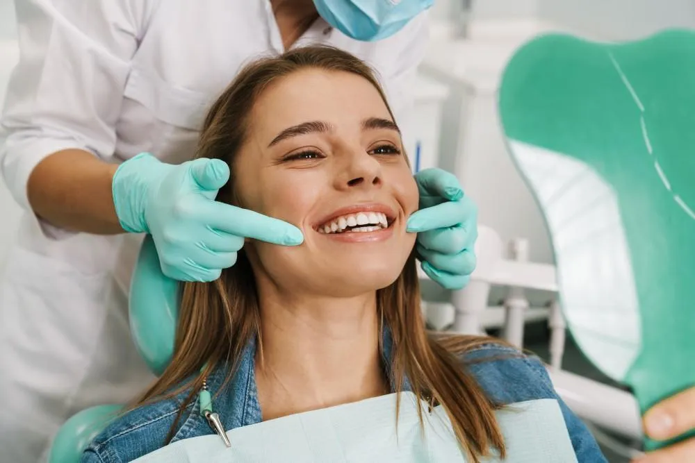 Smiling woman in a dental chair while a clinician in teal gloves gently pulls her cheeks to show her teeth during a checkup.
